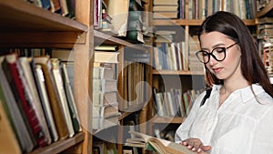 A young attractive student in glasses turns pages in the book, which she took from books on shelves in the library