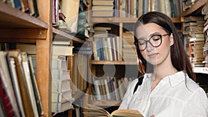 A young attractive student in glasses turns pages in the book, which she took from books on shelves in the library