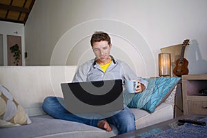 Young attractive and successful man working from home living room sitting on couch networking on laptop computer drinking coffee h