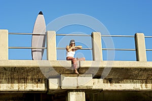 Young attractive surfer girl sitting on pier with surfboard