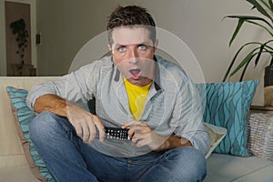 Young attractive surprised and shocked man in casual clothes at home with mouth opened in disbelief and surprise watching tv news