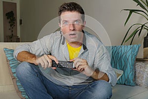 Young attractive surprised and shocked man in casual clothes at home with mouth opened in disbelief and surprise watching tv news