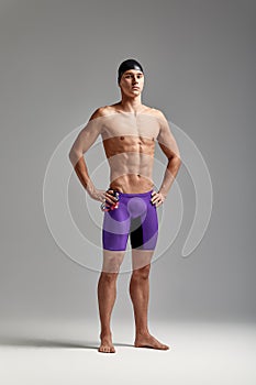 Young attractive swimmer in excellent physical shape in purple swimming shorts, on a gray background, copy space