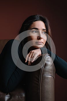 Young attractive thoughtful woman with long dark hair in black roll-neck sit on brown sofa putting head on crossed arms.
