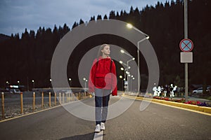 Young attractive tourist girl walking alone down the moutain highway in the evening. Female traveler in a red raincoat strolling