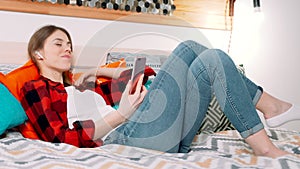 A young attractive white woman is lying on the bed and smiling in the modern bedroom while listening to musing using modern earbud