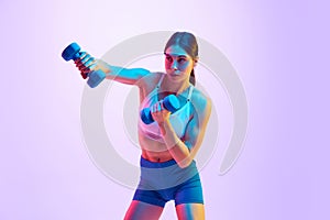 Young attractive woman in activewear boxing with dumbbells in hand in motion in neon light against gradient studio