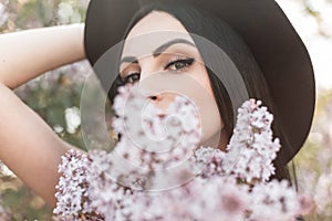 Young attractive woman with beautiful eyes in luxurious elegant hat covers face with bouquet fresh lilac flowers in flowering park