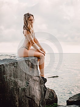 Young attractive woman in bikini posing at stones. Slim bikini girl at sea