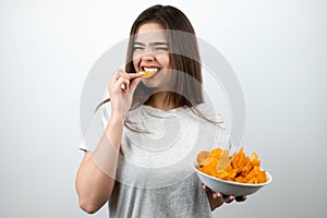 Young attractive woman biting with hunger one piece of chips in her hand standing with plate of spicy potatoe chips on isolated