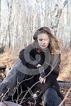 Young attractive woman in black coat sitting on the birch log in autumn forest