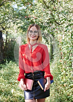 Young attractive woman with blond short hair, wearing red shirt and black skirt, holding books in her hands. Three-quarter