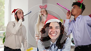 Young attractive woman blowing golden confetti from hands wearing christmas hat while her co-workers partying in the