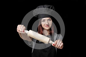 Young attractive woman chef in black uniform holds rolling pin on black background. Focus on rolling pin