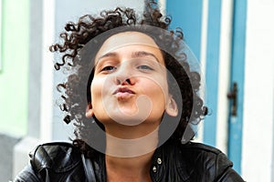 Young attractive woman with curly hair gesturing for a camera selfie and sending kisses. Urban city people concept