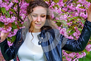Young attractive woman with curly hair in white dress and black leather jacket smiles in blossom of pink sakura flowers