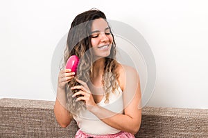 Young attractive woman with curly hairs brushing them while smiling on sofa at home