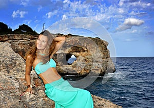 Young attractive woman on cyan glamour dress leaning on rock cliff at Spain coast