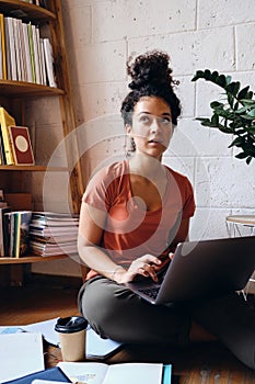 Young attractive woman with dark curly hair sitting on floor with laptop on knees thoughtfully looking aside with books