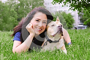 Young attractive woman with dark long hair is lying on the lawn with a dog, smiling and looking at the camera
