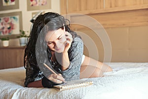 Young attractive woman designer smiling and drawing with pencil in notebook while lying on bed