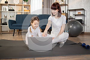 Young attractive woman doing sports gymnastics on the mat at home with her little cute daughter