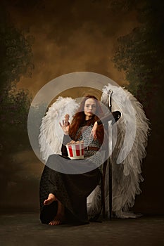 Young, attractive woman dressed as warrior-angel with large white wings sitting with box of snacks against vintage
