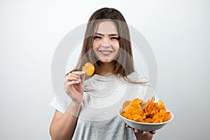 Young attractive woman holding one piece of chips in her hand standing with plate of spicy potatoe chips on isolated white