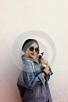 Young attractive woman hugging pussy cat in hands. Cute and glamorous girl in trendy sunglasses posing with her Siamese cat