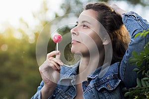 Young attractive woman licks caramel candy on an icicle on a stick in a park. A girl with a Chupa Chups in the open air.