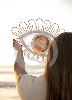 Young attractive woman lookig at herself on an eye-shaped mirror at golden sunset on the beach