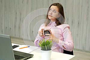 Young attractive woman at a modern office desk with coffee in hands, working with laptop, looking away, thinking about something