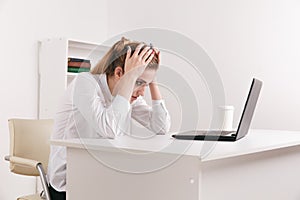 Young attractive woman at modern office desk, working on laptop, massaging temples to forget about constant headaches