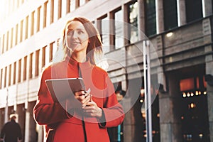 Young attractive woman in orange coat walks down city street holding tablet computer. Girl student going to lecture