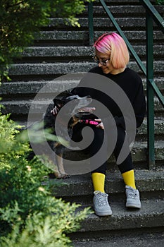 A young attractive woman with pink hair is sitting on the stairs in spring park with her Blue Heeler. Australian cattle