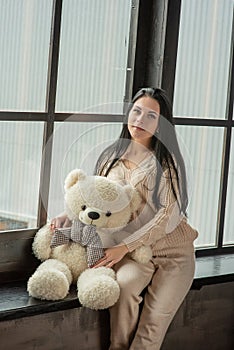 Young attractive woman posing. Girl at the window in a beige knitted suit. Girl with a soft plush toy bear in her hands