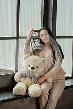 Young attractive woman posing. Girl at the window in a beige knitted suit. Girl with a soft plush toy bear in her hands