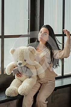 Young attractive woman posing. Girl at the window in a beige knitted suit. Girl with a soft plush toy bear in her hands