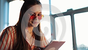 Young attractive woman in red sunglasses with tablet computer sitting in cafe. Beautiful girl in airport or shoppping