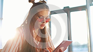 Young attractive woman in red sunglasses using tablet computer with touchscreen in a cafe. Beautiful girl in airport or