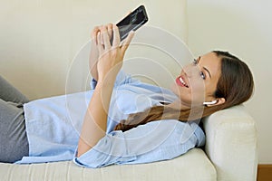 Young attractive woman relaxing on the couch at home choosing music on smart phone listening to music with wireless earphones