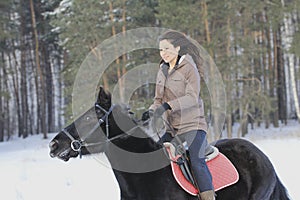 Young attractive woman riding on black horse in snowy forest