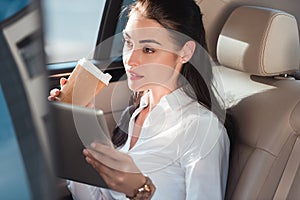 Young attractive woman sitting in a backseat of a car with digital tablet and cup