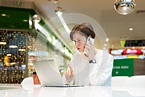 Young attractive woman sitting at a shopping center at a table and working at a computer laptop, using mobile phone