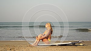 Young attractive woman is sitting on a surfboard. Beautiful woman surfer on the sandy beach against the background of