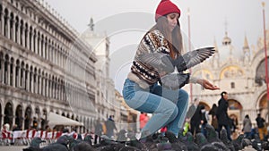 Young attractive woman squats in the square on the background of church and throws bread at a flock of pigeons