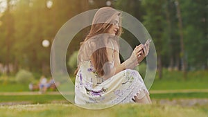 Young attractive woman in summer dress sits on green grass with mobile phone in hands. A park. Summer. Recreation.