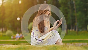 Young attractive woman in summer dress sits on green grass with mobile phone in hands. A park. Summer. Recreation.