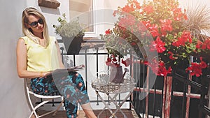 Young attractive woman with sunglasses enjoying sun and reading magazine on romantic balcony with flower boxes