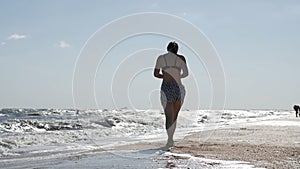 Young attractive woman in a swimsuit running on the sunny beach. Beautiful girl in sports uniform enjoys a walk along